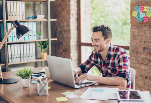 Cheerful young man smiling, typing on his laptop in nice modern work station at home, in casual smart wear