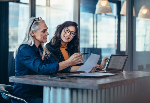 Two women analyzing documents while sitting on a table in office.