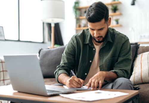 Focused smart indian or arabian guy, student or freelancer, sitting at home on sofa in living room, using laptop, working or studying remotely, listening to webinar, taking notes, homework
